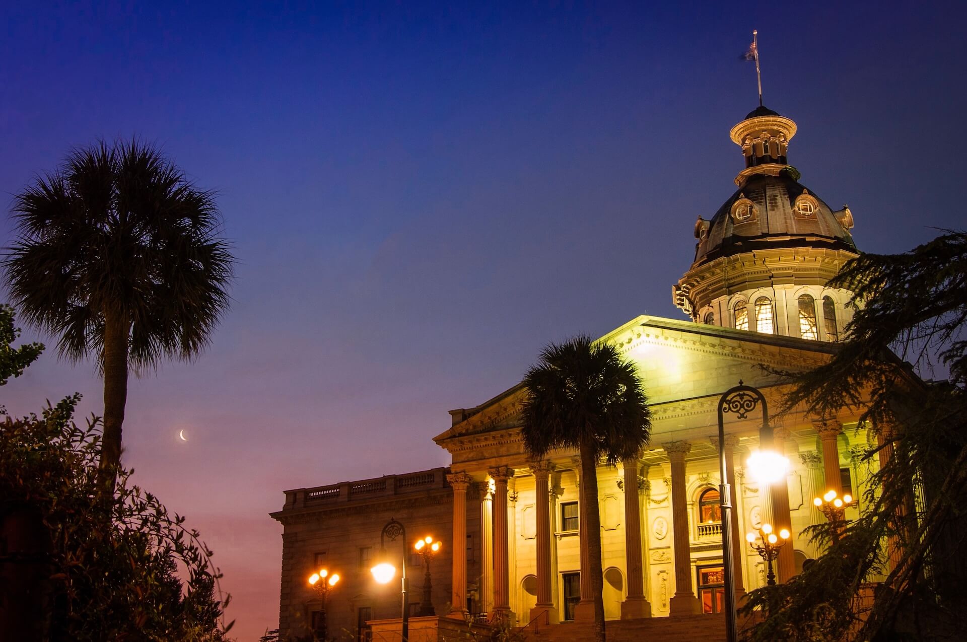 The image captures a historical, illuminated building with a dome and columns at dusk in South Carolina. Palm trees sway in the foreground while a crescent moon and streetlights hint at America’s future as high tech advances meet classic beauty in this serene evening scene.