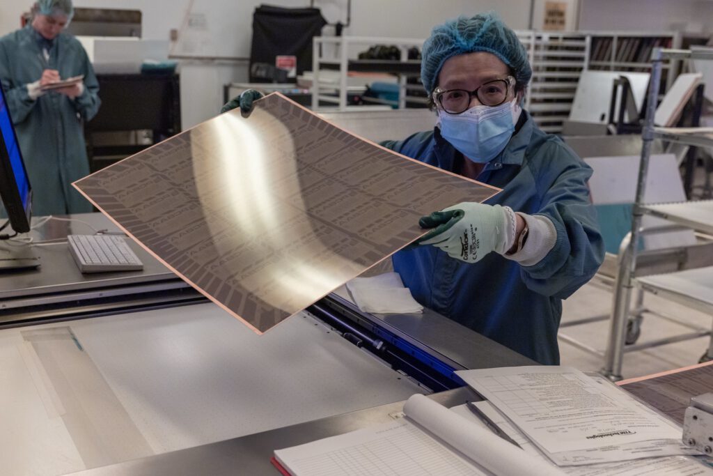 A person in protective clothing and a mask holds up a large sheet of thin, reflective material in a laboratory or manufacturing setting. Another person in the background is working on a computer, surrounded by equipment and documents.