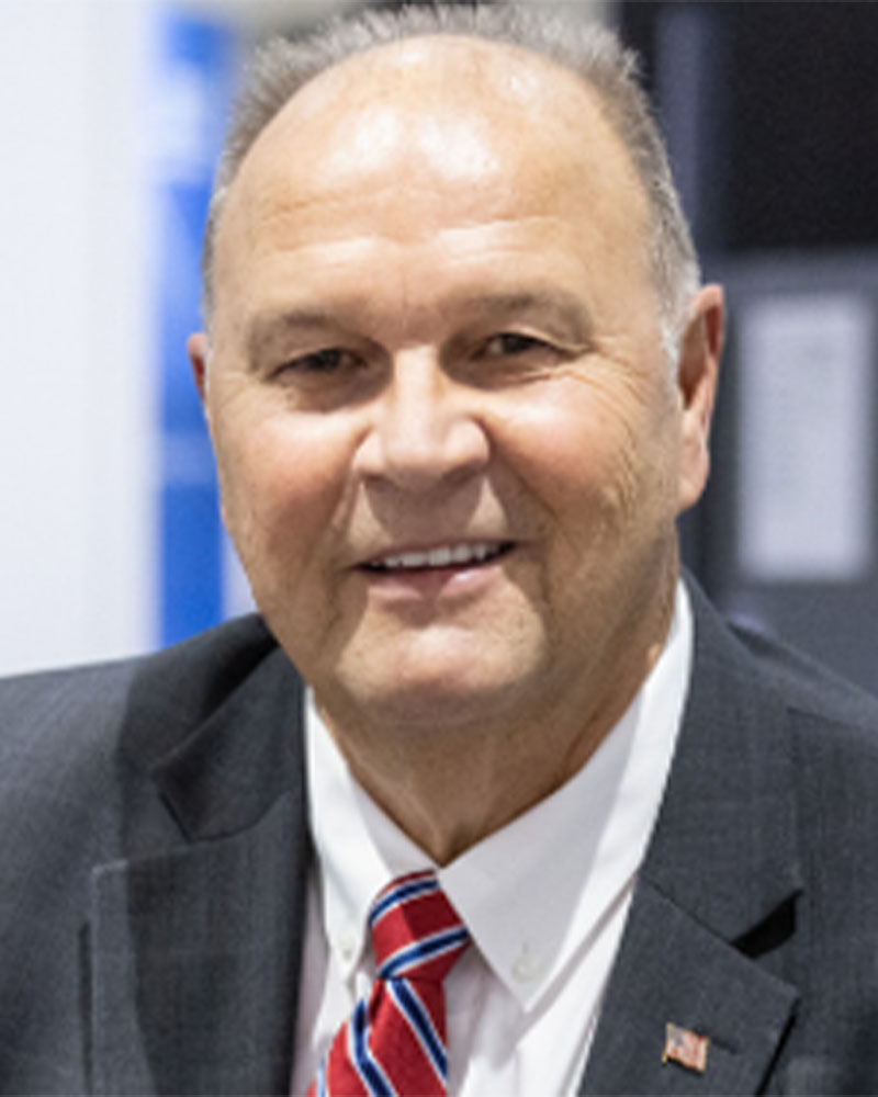 Shane Whiteside, a man in a suit and tie, is smiling at the camera. He wears a dark jacket with a small flag pin, complemented by a white shirt and red striped tie. The background is elegantly blurred.
